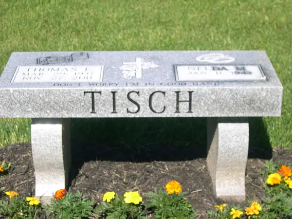 A light grey granite memorial bench with the surname cut large across the front, two name tablets above and a short epitaph between them
