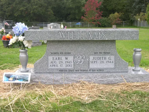 A grey granite memorial bench with the surname cut large across the seat edge, two name tablets set into the solid base, a carved emblem between them and matching granite vases
