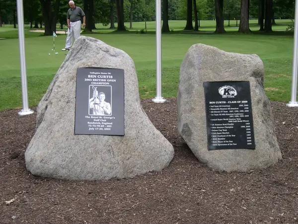 Two rough-hewn granite boulders on a golf course, each carrying a polished black granite plaque commemorating a British Open champion, with flagpoles behind