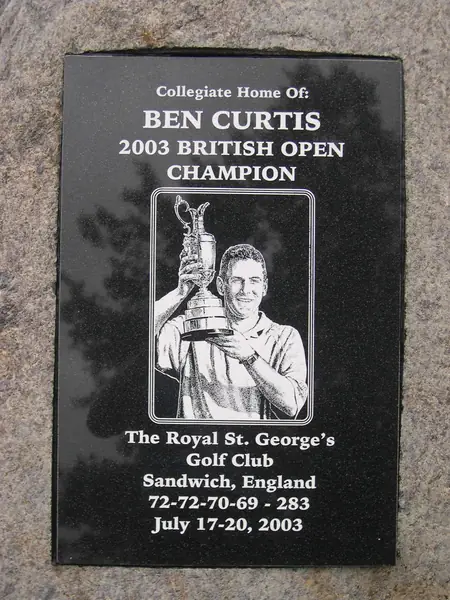 A polished black granite plaque set into a granite boulder, etched with a golfer lifting the Open trophy and the tournament details below