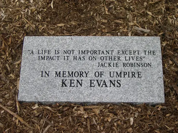 A grey granite memorial tablet set flush in mulch, carrying a quotation and a dedication to a baseball umpire