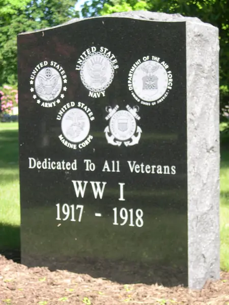 A polished black granite veterans memorial with a rock-pitched edge, carrying five service seals in relief above a First World War dedication and its dates