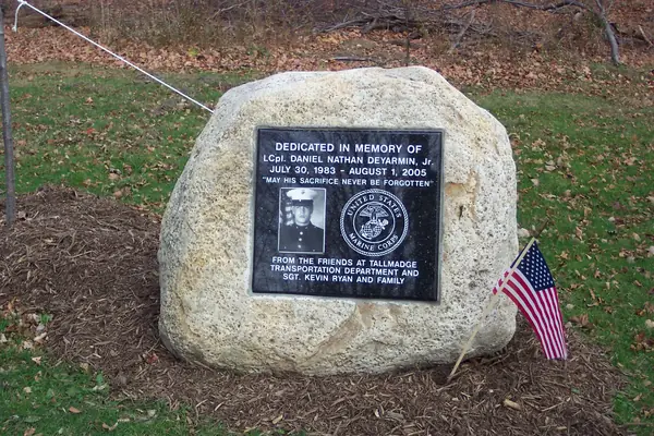 A large natural granite boulder carrying a polished black plaque with an etched portrait and a Marine Corps seal, dedicated to a fallen serviceman by a township transportation department