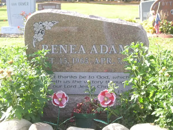 A grey granite monument with a rough-hewn top edge, carved flowers at one corner, the full name and dates on a single line and a scripture passage below