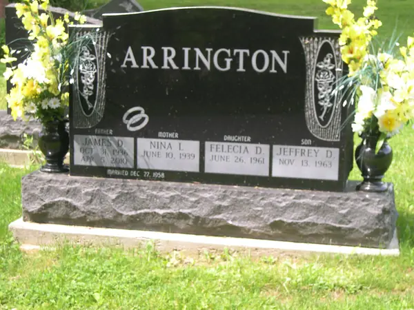 A polished black granite upright monument with ornamental carved panels at both shoulders and four name tablets labelled by relationship, on a rock-pitched base