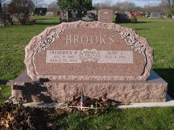 A rose granite monument with a scalloped top and carved floral corners, two name tablets and a marriage emblem between them, on a rock-pitched base