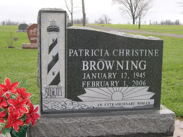 A two-piece monument pairing a tall grey granite pillar carved as a lighthouse with a dark granite tablet, etched with a sunrise over water and an epitaph