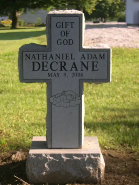 A grey granite monument cut in the shape of a Latin cross, carrying a short epitaph, a child's name and date, and a carved sleeping infant on clouds