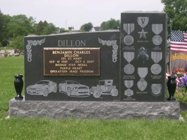 A polished black granite monument with a bronze service plaque, etched cars and a tall side panel of carved military medals and insignia