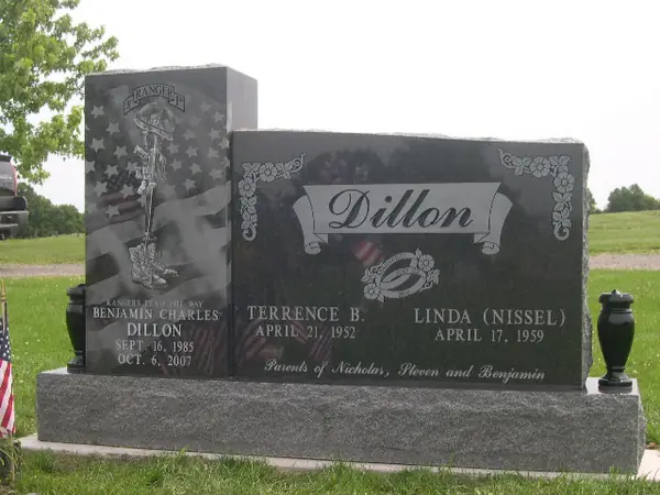 A two-part polished black granite memorial: a carved battlefield cross and helmet on the left panel and a family name in script on the right, with granite vases