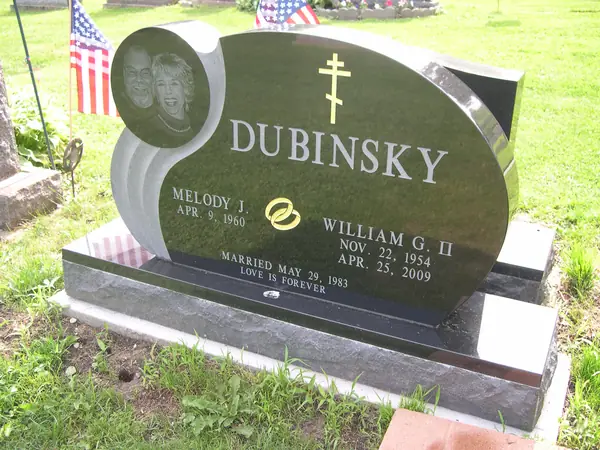 An oval polished green-black granite monument with an etched double portrait, an Orthodox cross, a wedding-ring emblem and a marriage date across the foot
