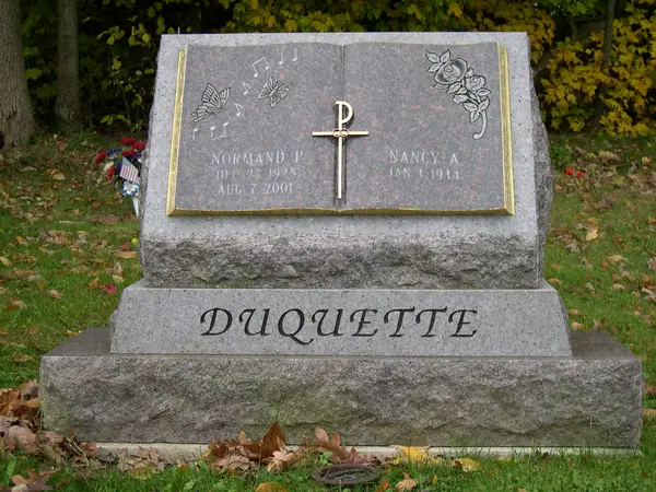 A grey granite monument built as a slanted open book on a stepped base, with a raised gilt cross, carved music and roses, and the family name cut into the base