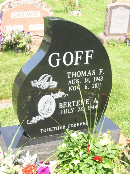 A polished black granite monument cut as a curved flame or leaf shape, with a wedding-ring emblem, praying hands and an epitaph across the foot
