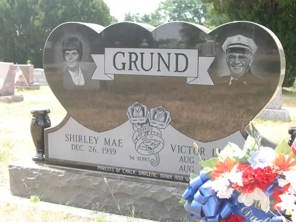 A polished granite monument cut as two joined hearts, each carrying an etched portrait, with a raised name panel, carved roses and a marriage banner