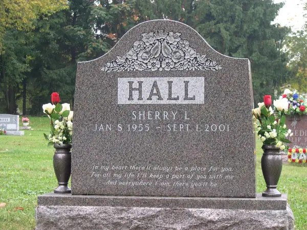 A grey granite monument with a shaped top, a carved floral swag, a raised name panel and a four-line epitaph in script, with bronze vases either side