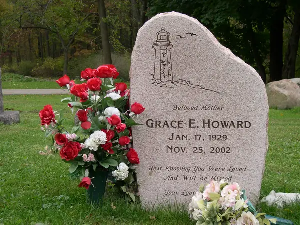 A grey granite monument with a rough-hewn edge, an etched lighthouse and gulls above a relationship line, the full name and dates, and an epitaph in script
