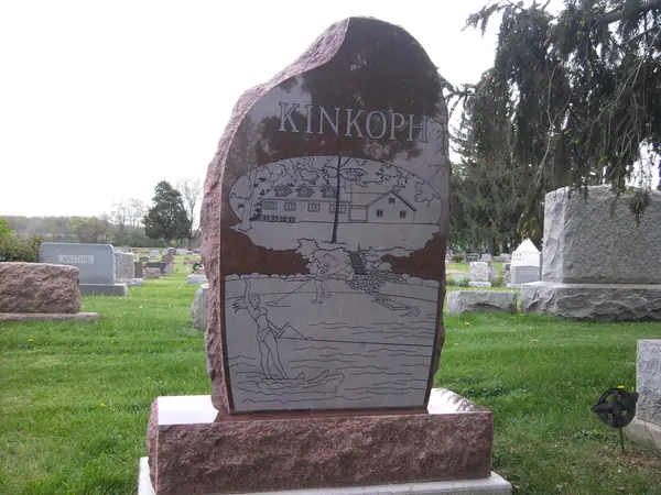 A red granite monument with a rough-hewn boulder profile, the family name across the top and an etched farm and waterfowl scene filling the face
