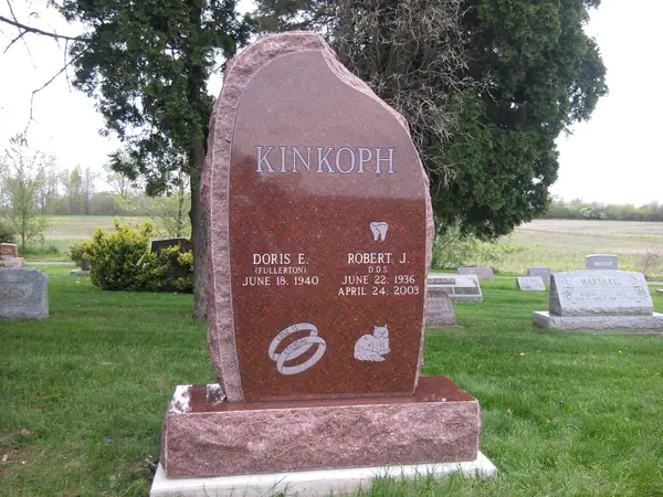 A red granite monument with a rough-hewn edge, the family name across the top, two name tablets and small carved emblems below