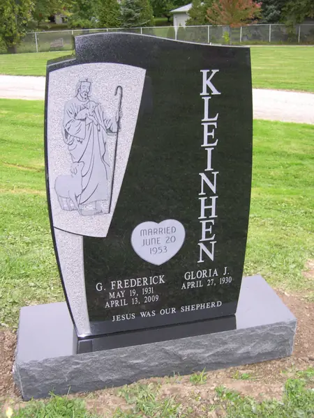 A polished black granite monument with a curved profile, a carved panel of Christ the Good Shepherd, the family name set vertically and a heart-shaped marriage plaque