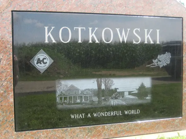 A polished black granite panel with the family name above an etched farm scene and a maker's emblem, with a short epitaph across the foot