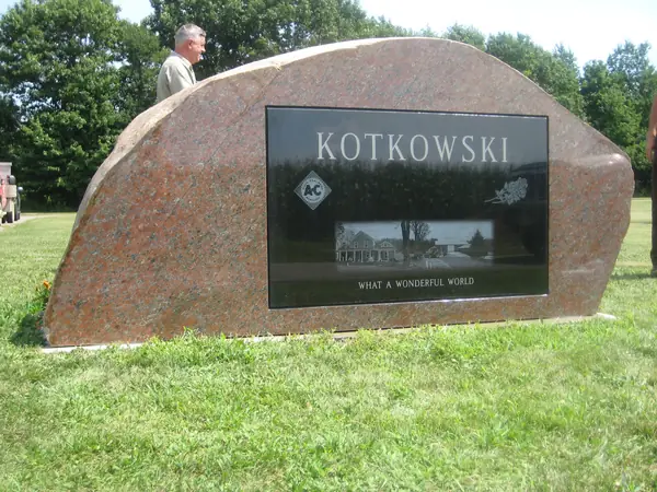A rose granite boulder-shaped monument with a large polished black granite panel set into its face, etched with a farmhouse scene and an epitaph