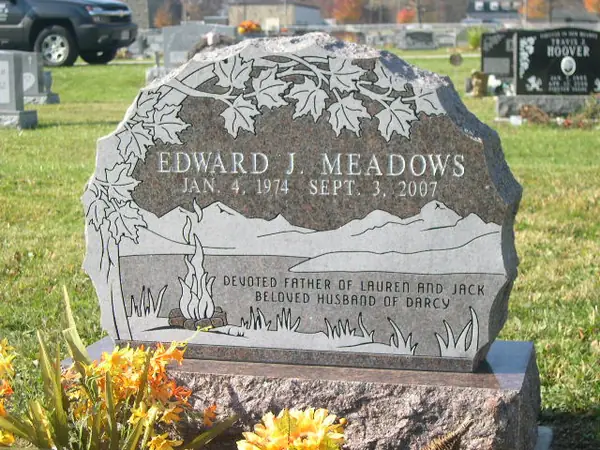 A grey granite monument with a rough-hewn boulder profile, a carved maple canopy across the top and an etched lake and cattail scene below the name