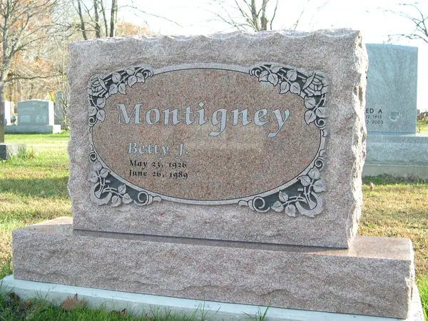 A rose granite monument with rough-hewn top and side edges, the family name in an ornamental oval frame carved with vines, and a single name below