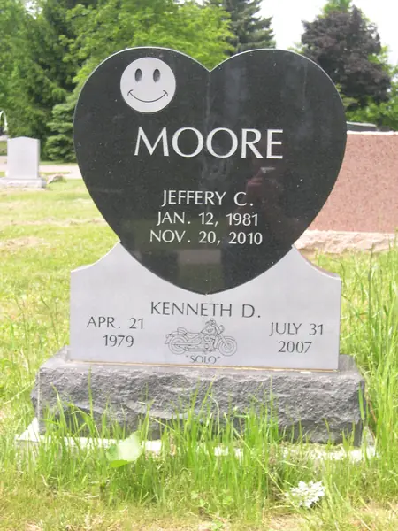 A monument built from a polished black granite heart, carved with a smiling face, above a light grey base carrying a second name and an etched motorcycle