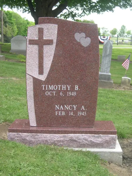 A red granite monument with a stepped, angled profile, a carved cross on a shield, two hearts and two names in cut lettering