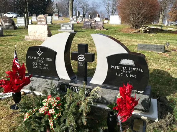 A polished black granite monument of two curved wings framing a freestanding cross, each wing carrying a name, with a Masonic emblem and an open book