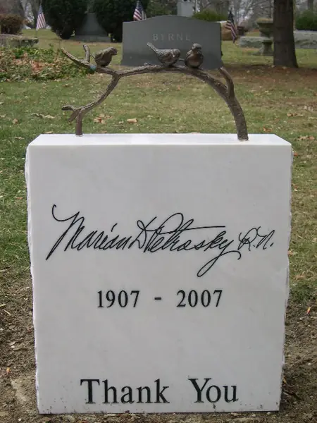 A white marble monument carrying a signature in script and a pair of dates, topped by a bronze branch with two perched birds