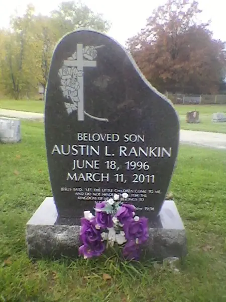 A polished black granite monument with a curved asymmetric profile, a carved cross with lilies, a relationship line and a scripture passage below the name