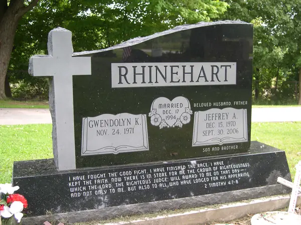A polished black granite monument with a wave-cut top and a full-height cross standing proud of the face, two names on open books and a long scripture passage on the base