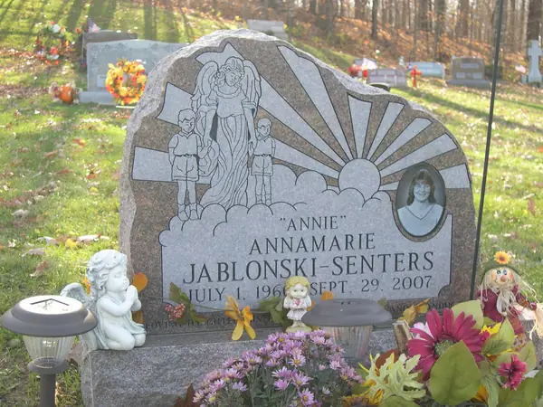 A grey granite monument with a rough-hewn edge, a carved scene of an angel with children against a sunburst, and an oval etched portrait beside the name
