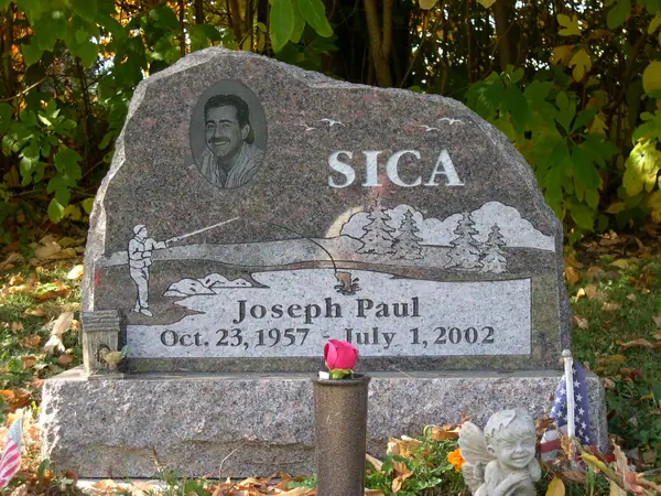 A grey granite monument with a rough-hewn boulder profile, an oval etched portrait and an etched fly-fishing and woodland scene beside the name
