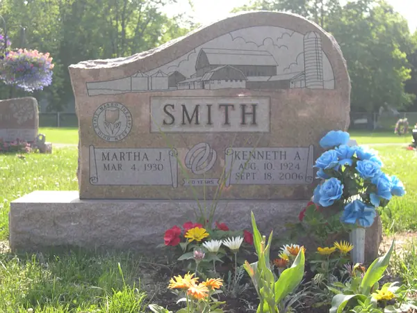 A grey granite monument with a wave-cut top, an etched barn and silo scene across the face, two name tablets and an anniversary emblem between them