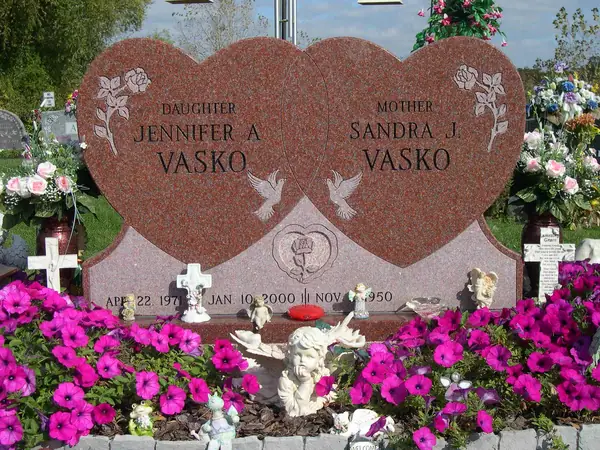 A red granite monument cut as two joined hearts, each carved with a rose and dove, carrying a relationship line above each name