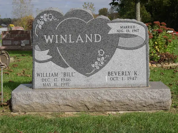 A grey granite monument cut as two overlapping hearts with a carved ribbon banner, floral sprays and a marriage date, above two name tablets