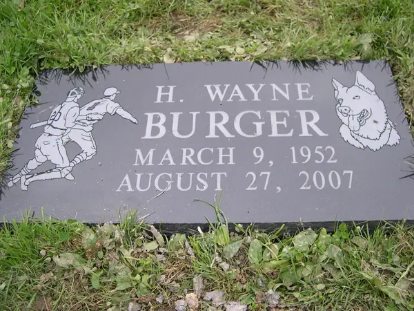 A flat dark granite marker with an etched baseball scene at one side and a terrier at the other, carrying a name and dates