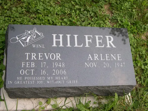 A grey granite bevel marker with an etched radio operator's emblem and callsign, two names and a two-line epitaph, on a rock-pitched base