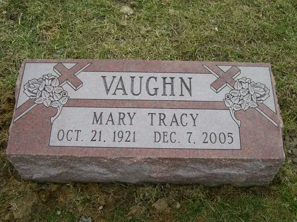 A red granite bevel marker with roses and crosses carved at both corners, a name and dates, on a rock-pitched base