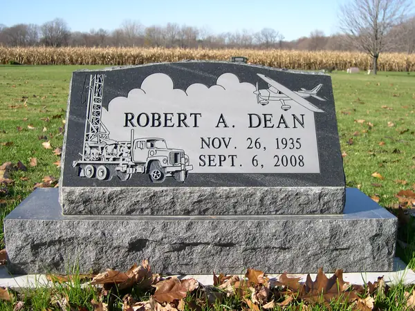 A polished dark granite slant marker with an etched drilling rig, tanker truck and aeroplane against clouds, and a name with dates