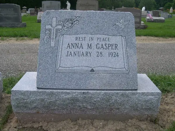 A light grey granite slant marker carved as an open book, with a cross, rose and dove above and a short epitaph with a name and date