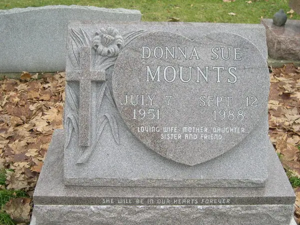 A grey granite slant marker with a large carved heart beside a carved cross and lilies, carrying a full name, dates and a relationship line