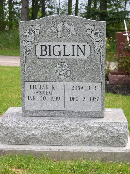 A light grey granite upright monument with a shaped top, roses carved at both shoulders and lovebirds above a raised name panel