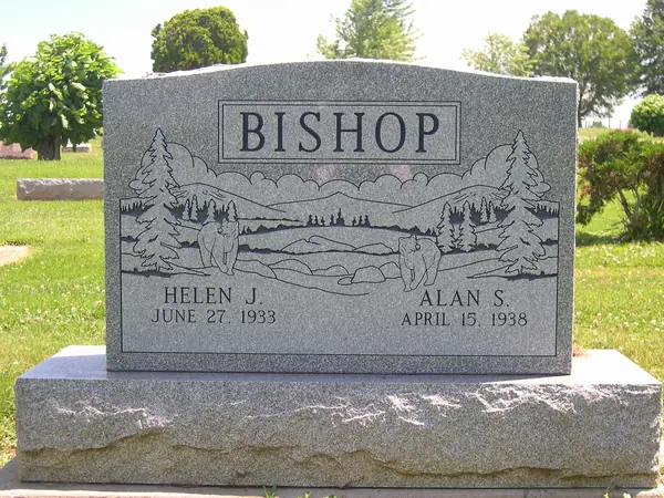 A grey granite upright monument with a broad etched woodland scene of deer, pines and mountains beneath a raised name panel, on a rock-pitched base