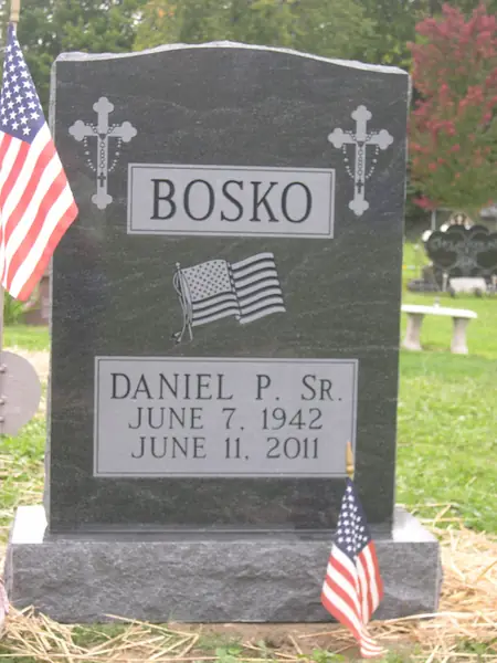 A grey granite upright monument with two carved crucifixes, a raised name panel and an etched American flag, with flags at the graveside