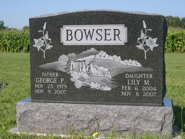 A polished black granite upright monument with a raised name panel, lilies carved at both shoulders and an etched mountain and waterfall scene across the face