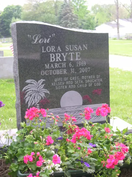 A dark granite upright monument with a wave-shaped top, a nickname in script above the full name, and a palm and sunset scene etched at the lower corner