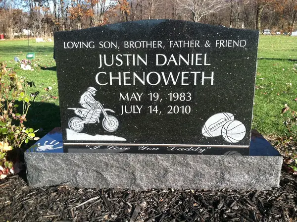 A polished black granite upright monument with a relationship line above the name, an etched motorcycle rider and a basketball, and an epitaph across the foot